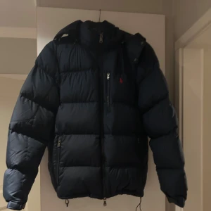 Mörkblå dunjacka från Polo Ralph Lauren - Säljer en mörkblå dunjacka från Polo Ralph Lauren med huva och dragkedja. Jackan har en klassisk pufferstil, flera fickor med dragkedja och den ikoniska röda loggan på bröstet. Perfekt för kalla dagar. Köpte förra vintern och har använts väldigt sparsamt 