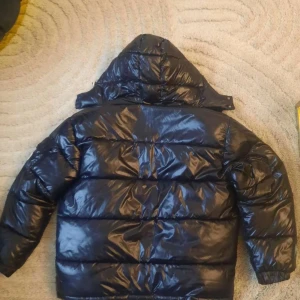 Svart Moncler pufferjacka med huva - Snygg svart pufferjacka från Moncler med glansig finish och klassisk quiltad design. Jackan har huva, dragkedja framtill och ikonisk Moncler-logga på ärmen. Insidan har ett coolt serietidningsmärke. Perfekt för kalla dagar när du vill hålla stilen.