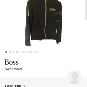 Svart hoodie med guld-detaljer från Boss - Svart hoodie från Boss med dragkedja i guld och matchande guldlogga på bröstet. Tröjan har huva, långa ärmar och fickor framtill. Snygga detaljer med guldfärgade dragkedjor även på ärmen. Perfekt för en stilren och trendig look. Skriv vid minsta fundering! 