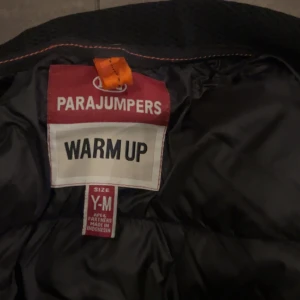 Svart pufferjacka Parajumpers Y-M - Svart pufferjacka från Parajumpers med quiltad framsida och rygg, klassisk gul detalj vid kragen och patch på ärmen. Jackan har dragkedja framtill och är tillverkad i syntetmaterial. Perfekt för kyliga dagar och har en clean, sportig look.