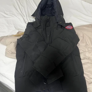 Canada Goose Wyndham - Säljer en svart dunjacka från Canada Goose med huva och klassisk logga på ärmen. Jackan är tjock och varm, perfekt för kalla vinterdagar. Den har dragkedja och knappar framtill samt flera fickor. Snygg och stilren design som håller dig varm hela vintern. Den är i väldigt bra skick då jag använt den fåtal gånger och har inga defekter.