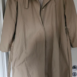Beige trenchcoat från Stockh LM, stl 44 - Klassisk beige trenchcoat från Stockh LM i storlek 44. Jackan har knäppning framtill, bälte i midjan och långa ärmar. Snygg och tidlös modell med slät yta och enkel design, perfekt för dig som gillar stilrena plagg.
