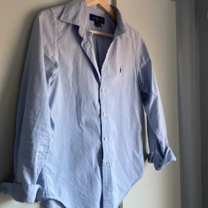 Ljusblå skjorta från Ralph Lauren - Klassisk ljusblå skjorta från Ralph Lauren med broderad logga på bröstet. Skjortan har lång ärm, knappar framtill och en snygg krage. Perfekt för dig som gillar stilrena och tidlösa plagg. Tillverkad i mjuk bomull för en skön känsla.
