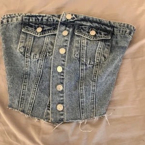 Denim tubtopp med knappar - Cool tubtopp i blå denim med knappar framtill och råa, fransiga kanter. Toppen har två bröstfickor med lock och knappdetaljer samt smockad baksida för extra stretch. Perfekt för dig som vill sticka ut med en edgy jeanslook.