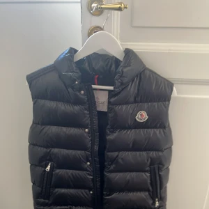 Moncler Svart dunväst - Säljer en Grisch svart Moncler väst, quiltad dunväst från Moncler med hög krage och dragkedja framtill. Västen har två fickor med dragkedja och klassisk Moncler-logga på bröstet. Insidan har ett färgglatt serietryck. Perfekt för lager-på-lager och snygg till streetwear.