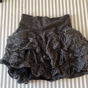  kortkjol från Sofie Schnoor - Supersnygg kortkjol från Sofie Schnoor i mörkgrått med volangdetaljer och stretchig midja. Kjolen har ett coolt, rynkat tyg med massor av glitter som ger extra attityd. Perfekt för dig som vill sticka ut med en unik look.