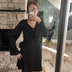 Svart omlottklänning från NA-KD - Supersnygg svart omlottklänning från NA-KD i glansigt satintyg. Klänningen har långa vida ärmar, knyt i midjan och v-ringning. Perfekt för dig som gillar en stilren och trendig look med lite extra edge.