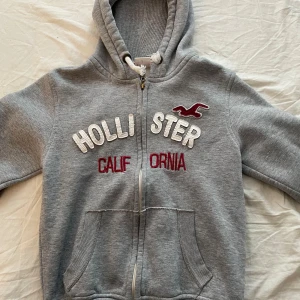 Grå hoodie från Hollister med brodyr - Grå hoodie från Hollister med vit och röda detaljer. Den har några defekter på insidan av märket men inget som förstör trycket på utsidan. 