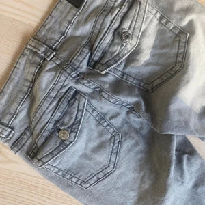 Grå jeansbyxor med stora fickor - Snygga grå jeansbyxor från Perfect Jeans med stora fickor med lock och knapp bak. Klassisk femficksmodell med bälteshällor och silverfärgade knappar. Perfekt för dig som gillar en avslappnad och cool stil.