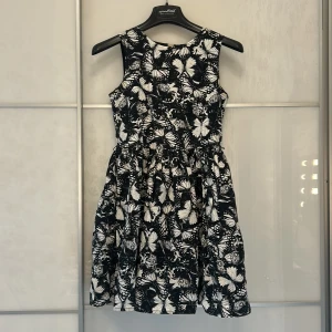 Svartvit ärmlös klänning med fjärilar - Svartvit kortklänning från H&M med ett fjärilsmönster. Klänningen är ärmlös och har en rund halsringning samt en markerad midja. Baksidan har en dragkedja för enkel påklädning. Perfekt för dig som gillar unika prints!