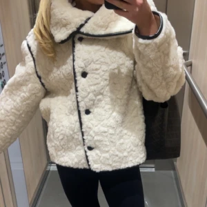 Vit teddyjacka från Gina Tricot - Supermysig vit teddyjacka från Gina Tricot med svarta detaljer längs kanterna och stora knappar framtill. Jackan har en oversized krage som man kan knäppa längst upp. Säljer för att den inte kommer till användning 