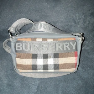 Rutig axelväska från Burberry - Snygg axelväska från Burberry med klassiskt rutigt mönster i beige, brun, svart, vit och röd. Väskan har bred grå axelrem med Burberry-logga, detaljer i grått och dragkedja med färgglad snörning. Perfekt storlek för det viktigaste och har innerfack med dragkedja.