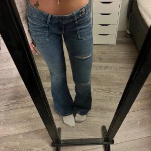 Blå bootcut jeans med slitningar - Snygga blå jeans med bootcut passform och slitna detaljer på låren. Jeansen har stora fickor fram och klassisk knappstängning. Aldrig använd!