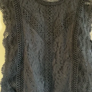 Svart spetsblus från ONLY - Supersnygg svart blus från ONLY med detaljerad spets och vackra vågiga kanter. Blusen är ärmlös och har ett feminint mönster i spetsen som ger en elegant look. Perfekt att styla med jeans eller kjol för en trendig outfit.