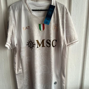 Vit Napoli fotbollströja EA7 De Bruyne - Napoli fotbollströja i vitt från EA7 med De Bruyne och nummer 11 på ryggen. Tröjan har korta ärmar, guldiga och svarta detaljer, diskret mönster och klubbmärke på bröstet. Tillverkad i lätt och ventilerande material, perfekt för match eller träning.