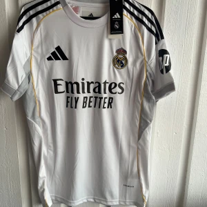 Real Madrid Mastantuono matchtröja - Säljer en officiell Real Madrid fotbollströja från Adidas med Mastantuono och nummer 30 på ryggen. Tröjan är vit med svarta detaljer, gula linjer och klubbmärke på bröstet. Kortärmad modell i lätt funktionsmaterial med broderade loggor och tryckta sponsorer.