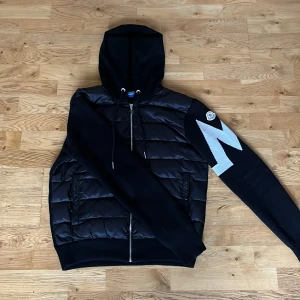 Svart pufferjacka från Moncler - Svart pufferjacka från Moncler med huva och dragkedja framtill. Jackan har quiltade partier på framsidan, släta ärmar och en vit blixtdetalj på ena ärmen. Två fickor med knapp och ribbade muddar. Säljes för 2100 då skickat är perfekt men kan diskuteras. Storleken är L men passar M och S