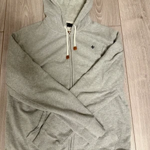 Grå hoodie från Morris med dragkedja - Säljer en ljusgrå hoodie från Morris med dragkedja och huva. Tröjan har vita snören med bruna detaljer, ribbade muddar och en liten broderad logga på bröstet. Perfekt för chill dagar och enkel att matcha med jeans eller joggers.