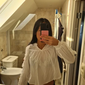 Vit blus med volangdetaljer - Supersöt vit blus från Gina tricot Young med volangdetaljer längs ärmarna och smock vid ärmsluten. Blusen har en lös passform, v-ringad hals och långa ärmar. Perfekt för dig som gillar romantisk och chill stil. Materialet är mjuk bomull.