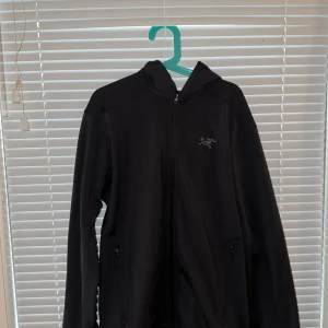 Svart från Arc'teryx  - Svart fleecejacka från Arc'teryx med huva och dragkedja framtill. Jackan har två sidofickor med dragkedja och diskret logga på bröstet. Perfekt för lager-på-lager och outdoor. Enkel, stilren design med lång ärm.aldrig använd btw!!