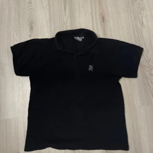 Svart pikétröja från Millbrooke S - Klassisk svart pikétröja från Millbrooke i storlek S. Tröjan har korta ärmar, krage och knäppning framtill samt en broderad lejonlogga på bröstet. Perfekt för en clean och sportig stil.