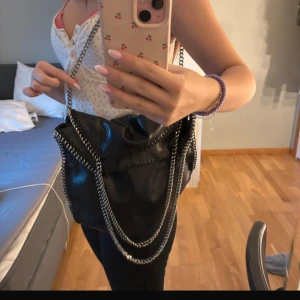 Svart axelväska Stella McCartney - Säljer en svart axelväska från Stella McCartney med snygga silverfärgade kedjor som axelrem och runt kanterna. Väskan är i veganskt skinn och har ett coolt innerfoder med logotypmönster. Perfekt för dig som vill ha en trendig och rymlig väska.