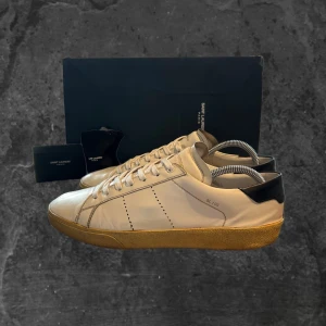 Saint Laurent SL/06 vita sneakers - Saint Laurent SL/06 sneakers i vitt skinn med beige sula och svarta detaljer vid hälen. Klassisk låg modell med snörning och diskreta perforerade linjer på sidorna. Märkeslogga på plösen och hälen. Perfekta för en clean och stilren look.