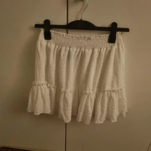 Vit volangkjol från SHEIN - Supersöt vit kortkjol från SHEIN med volangdetaljer och smockad midja. Kjolen har en lätt textur i tyget och är perfekt för dig som gillar en feminin och luftig stil. Passar dig som vill ha en enkel men ändå trendig look.