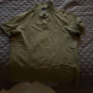 Olivgrön pikétröja från Gant XL - Snygg olivgrön pikétröja från Gant i klassisk modell med krage och knappar framtill. Tröjan har korta ärmar och en liten broderad logga på bröstet. Perfekt för en avslappnad men stilren look.