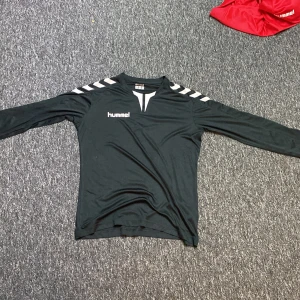 Svart långärmad fotbollströja Hummel - Svart långärmad fotbollströja från Hummel med vita detaljer på axlarna och logga på bröstet. Tröjan har en sportig design med rund hals och är tillverkad i ett lätt och funktionellt material som andas. Perfekt för träning eller match.