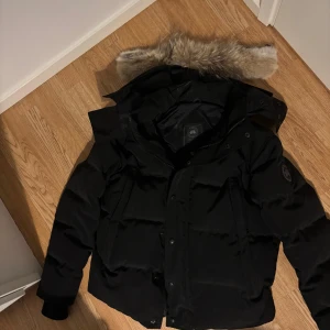 Svart dunjacka från Canada Goose M - Säljer en svart dunjacka från Canada Goose i storlek M. Jackan har en fluffig pälskant på huvan, black label  på ärmen och dragkedja med knappar framtill. Perfekt för kalla dagar och riktigt snygg streetstil.