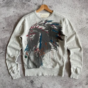 Grå sweatshirt med färgstarkt tryck från denim and supply( intressekoll) - Säljer en grå sweatshirt från denim and supply med ett stort, färgstarkt tryck framtill i svart, blått och rött. Tröjan har rund hals, ribbade muddar och är i mjuk bomull. Perfekt för dig som gillar unika prints och streetstyle.(två små hål som visas på bild)