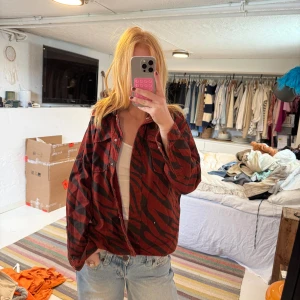 Röd zebramönstrad overshirt - Säljer en oversized röd overshirt med svart zebramönster. Skjortan har klassisk krage, knäppning framtill och långa ärmar. Perfekt att slänga över en t-shirt för en cool och avslappnad look.
