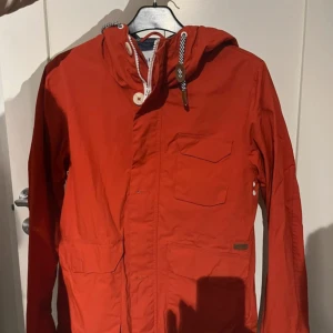 Röd vindjacka från Elvine med huva - Säljer en röd vindjacka från Elvine i storlek S. Jackan har huva med snörning, flera fickor framtill och snygga knappar. Perfekt för blåsiga dagar och har en sportig vibe. Insidan är fodrad med mesh för extra komfort.