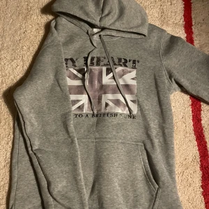 Grå hoodie med brittisk flagga - Grå hoodie med tryck av en sliten brittisk flagga och texten 'MY HEART BEATS TO A BRITISH TUNE' på bröstet. Tröjan har huva med snörning och en stor magficka. Perfekt för dig som gillar streetstyle och vill sticka ut med en cool print.(funkar som xs)