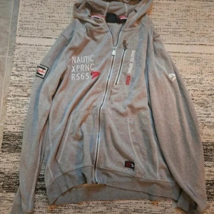 Grå hoodie med dragkedja Nautic XPRNC - Säljer en grå hoodie från Nautic XPRNC med hel dragkedja, huva och tryck på bröstet och ärmen. Tröjan har praktiska fickor framtill och detaljer i vitt och rött. Perfekt för dig som gillar sportig och avslappnad stil.