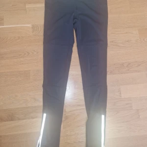 Svarta träningstights från Saint Tropez - Svarta träningstights från Saint Tropez med reflekterande detaljer vid bensluten. Insidan har en grön och grå kontrastfärg. Tightsen är tighta och gjorda i ett stretchigt syntetmaterial, perfekta för träning och löpning.