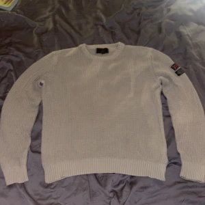 Beige stickad tröja Morris - Säljer en beige stickad tröja från Morris med rund hals och ribbade muddar. Tröjan har en snygg patch med brittisk flagga och text på ena ärmen. Perfekt för dig som gillar klassisk och stilren look. SKRIV VID MINSTA FRÅGA, PRIS KAN DISKUTERAS.