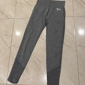 Grå leggings från RW med hög midja - Säljer ett par grå leggings från RW med hög midja och snygga stickade detaljer längs benen. De är tighta och har en diskret logga framtill. Perfekta för träning eller chill dagar. Materialet är mjukt och stretchigt syntet för maximal komfort.