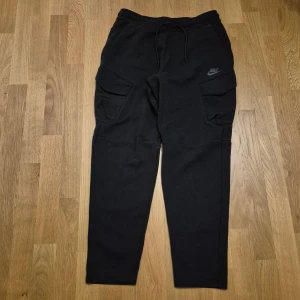 Svarta cargopants från Nike - Svarta cargopants från Nike med stora fickor på sidorna och dragsko i midjan. Byxorna har en avslappnad passform och är tillverkade i mjukt bomullsmaterial. Perfekta för en chill och sportig stil. Nike-logga tryckt på ena benet.