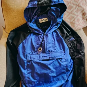 Lundhags vindjacka blå/svart stl small - Säljer en blå och svart vindjacka från Lundhags med huva och stor magficka. Jackan har half zip framtill, dragsko i huvan och broderad logga på ärmen. Perfekt för dig som gillar outdoor och vill ha en snygg och funktionell jacka.