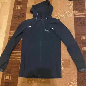 Svart vindjacka från Under Armour - Svart vindjacka från Under Armour med huva och hel dragkedja framtill. Jackan har reflekterande detaljer och logga på bröstet. Långärmad modell med sportig look, perfekt för utomhusaktiviteter.