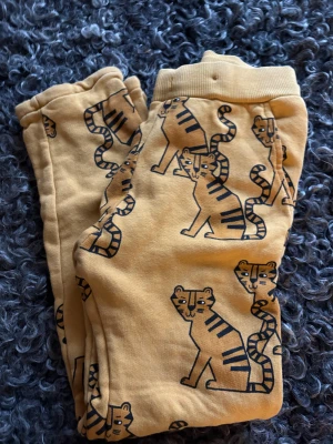 Gula mjukisbyxor med tigerprint Lindex - Säljer ett par mjukisbyxor från Lindex i storlek 122. Byxorna är senapsgula med coolt tigerprint över hela och har ribbad midja. Sköna och mjuka, perfekta för chill dagar. Materialet är bomull och de har normal passform.