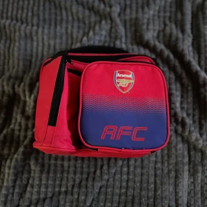 Arsenal röd och blå lunch box - Snygg lunch box i rött och blått med Arsenal-logga och AFC-tryck på framsidan. Väskan har flera fack med dragkedja och är tillverkad i slitstark polyester. Perfekt för dig som vill visa ditt stöd för Arsenal och ha plats för allt du behöver. Den är i jättebra skick och passar för dig som behöver matlådor osv i vardagen. Hör av er om ni har frågor!
