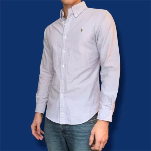 Ljusblå skjorta från Ralph Lauren - Snygg ljusblå skjorta från Ralph Lauren med klassisk button-down krage och broderad logga på bröstet. Skjortan är tillverkad i 100% bomull och har lång ärm samt vita knappar framtill. | Storlek M | Bröstmått 50 cm | Längd 70 cm | 