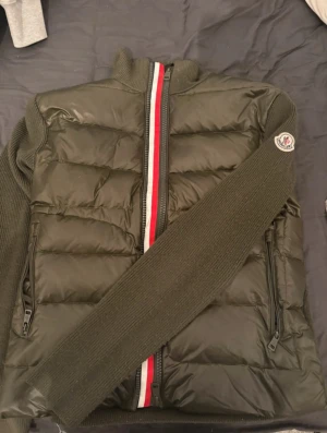 Moncler mörkgrön cardigan  - Snygg mörkgrön cardigan från Moncler med stickade ärmar och rygg. Jackan har quiltad framsida, dragkedja med röd och vit rand, samt Moncler-logga på ärmen. Perfekt för dig som vill ha en stilren och varm jacka med coola detaljer.