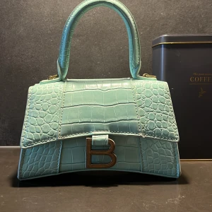 Mintgrön handväska i skinn - Säljer en mintgrön handväska med spänne, krokodilpräglat skinn och guldfärgade metalldetaljer. Väskan har en avtagbar axelrem. Vid frågor, hör gärna av dig! 