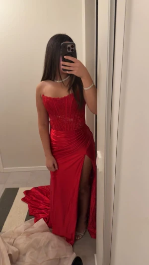 Röd långklänning med spets och slits - Säljer en röd axelbandslös långklänning med spetsdetaljer upptill och markerad midja. Kjolen är i glansigt tyg med hög slits på ena sidan och ett elegant släp. Perfekt för bal eller fest. Sitter tight och har en snygg omlott-effekt vid höften. Endast andvänd en gång