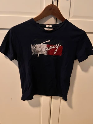 Mörkblå Tommy Hilfiger t-shirt - Snygg mörkblå t-shirt från Tommy Hilfiger med stort tryck på bröstet i vitt och rött. Klassisk rund hals och korta ärmar. Materialet är mjuk bomull som känns skönt mot huden. Perfekt för dig som gillar stilrena logotoplagg.