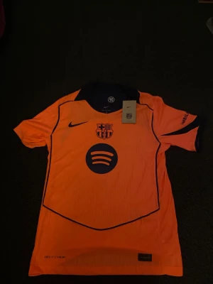 Barcelona Raphinha 11 orange matchtröja - Barcelona fotbollströja med Raphinha och nummer 11 på ryggen. Tröjan är orange med mörkblå detaljer, klubbmärke på bröstet och sponsorloggor från Spotify och UNHCR. Kortärmad, tillverkad i lätt Dri-FIT material för maximal komfort på planen.